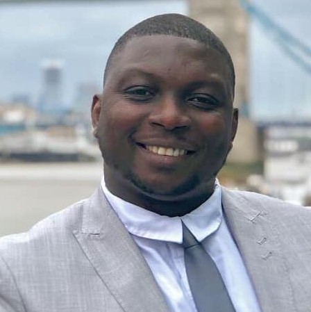 Dr Daniel Ogidigben smiles wearing a grey suit outside on a cloudy day.