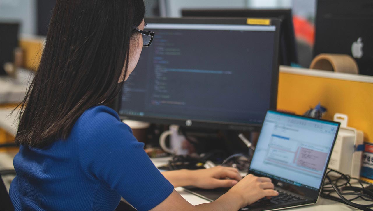 A woman with her back to the camera, dressed in blue, works on a laptop at her desk.