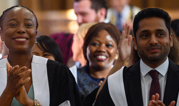 Two women and a man wearing ceremonial robes stand up to clap at something or someone out of shot.