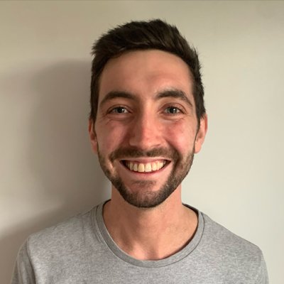 Dr Tom France smiles at the camera while standing in front of a cream wall. 