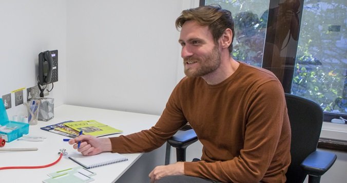 A doctor in a brown jumper sitting and a desk and smiling at someone off-camera while taking notes