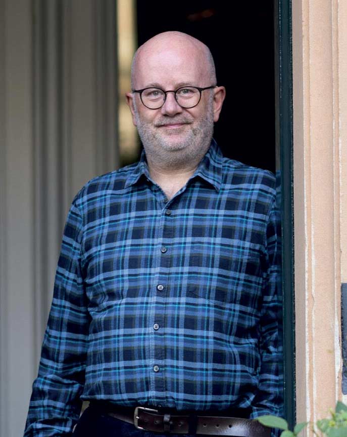 A portrait photo of Dr Ewen Stewart standing in a doorway wearing a blue tartan shirt. 
