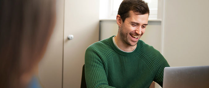 Man wearing a green jumper, smiling whilst looking at a laptop screen. A colleague is sat next to him.