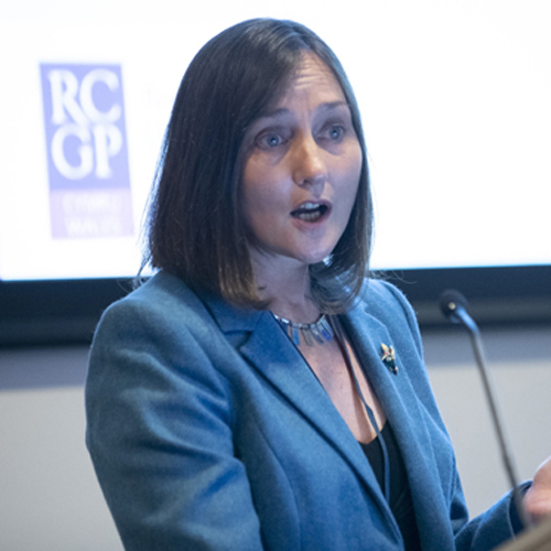 A square photograph of Dr Rowena Christmas at a lectern delivering a speech in front of a large screen.