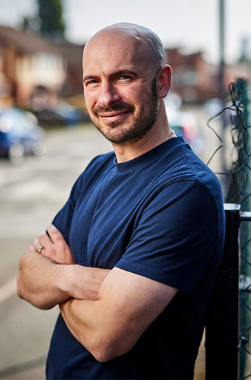 Dr Benjamin Brown leans against a wire fence with a street in the background.