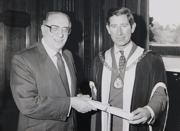 Black and white image of Prince Charles in ceremonial wear, being handed a scroll by a man in a suit.