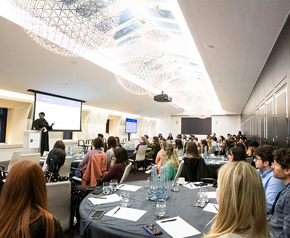 A square image of attendees sitting around circular tables at an event.