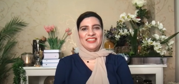 A woman in a headscarf smiles at the camera. She is sitting in front of a desk adorned with pink and white flowers, and a pile of books.