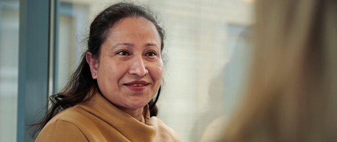 A woman with dark hair wearing a beige jumper smiles at someone out of shot.