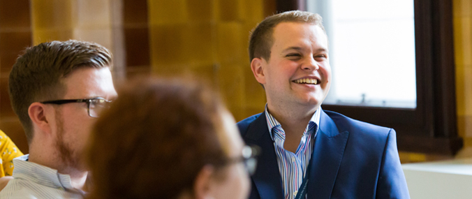 A smiling man wearing a navy suit sits with two colleagues.
