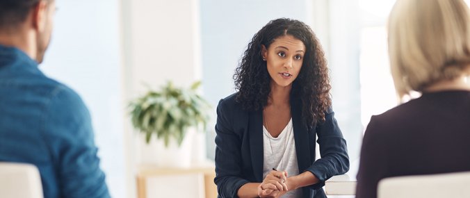 A seated woman wearing a black jacket facing forward sits between two seated patients facing away from the camera.
