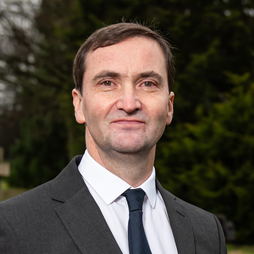 A square image of Dr Chris Provan standing against a cream wall wearing a shirt and tie.