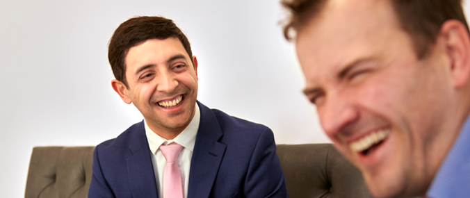 A man with dark, cropped hair looks at a laughing colleague close to the camera. 