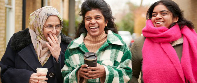 Three women laughing and smiling, all wearing coats and scarves. Two women are holding coffee cups.